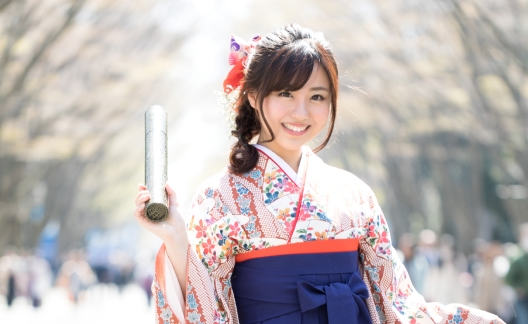 桜並木を背に片手で卒業証書の筒を手にしている袴姿の女学生