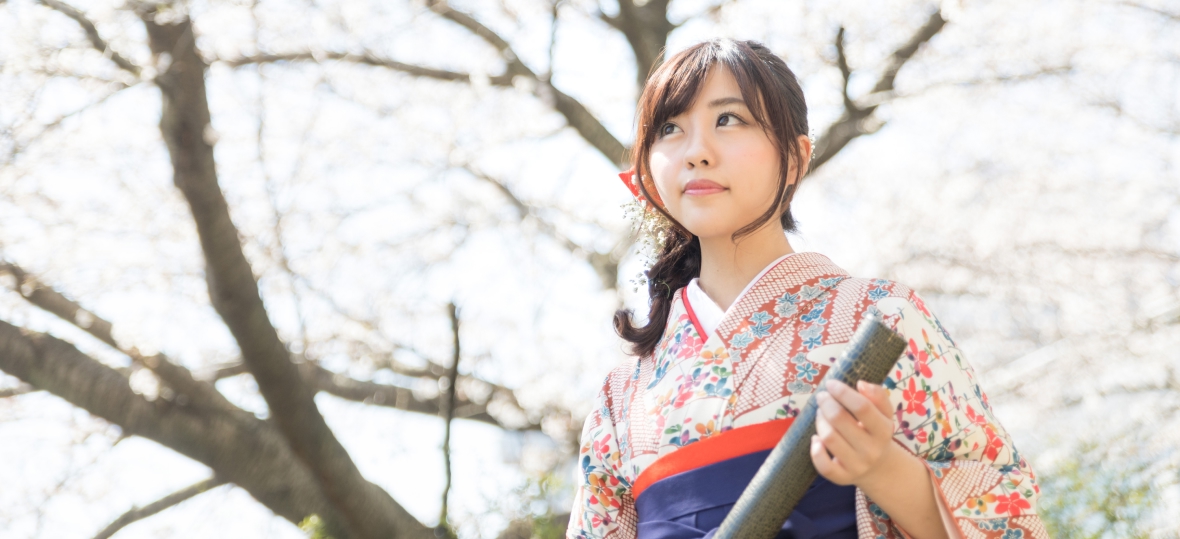 桜の木をバックに卒業証書を持つ袴姿の女学生