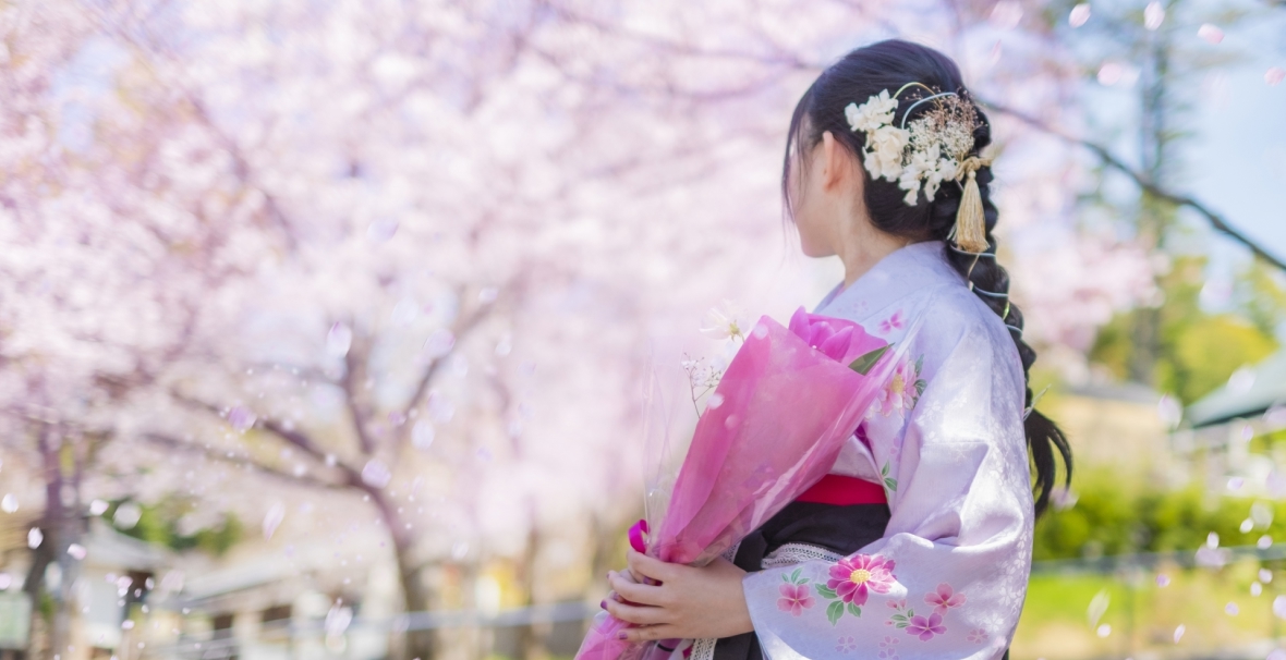 薄いピンクの花柄の着物の袴姿の女性が花束を持ち桜の花を振り返り見ている